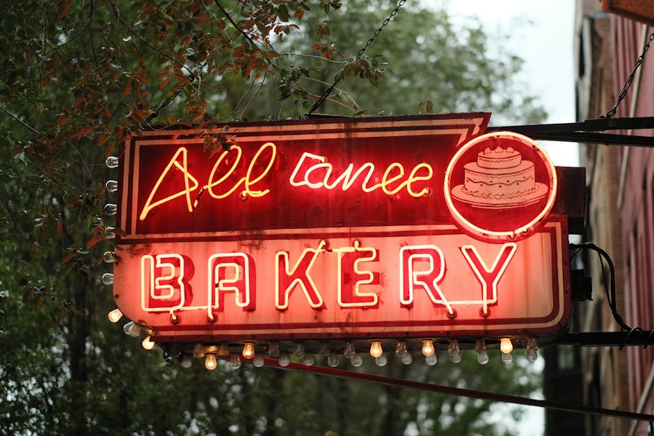 Retro neon bakery sign featuring cake icon on a tree-lined urban street.